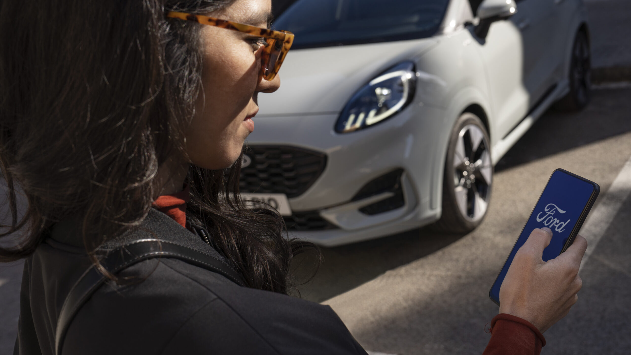 Person hält Smartphone mit Ford-App vor einem geparkten weißen Auto